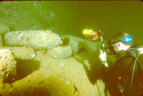 Scuba diver holding a camera toward stacked shells inside a shipwreck